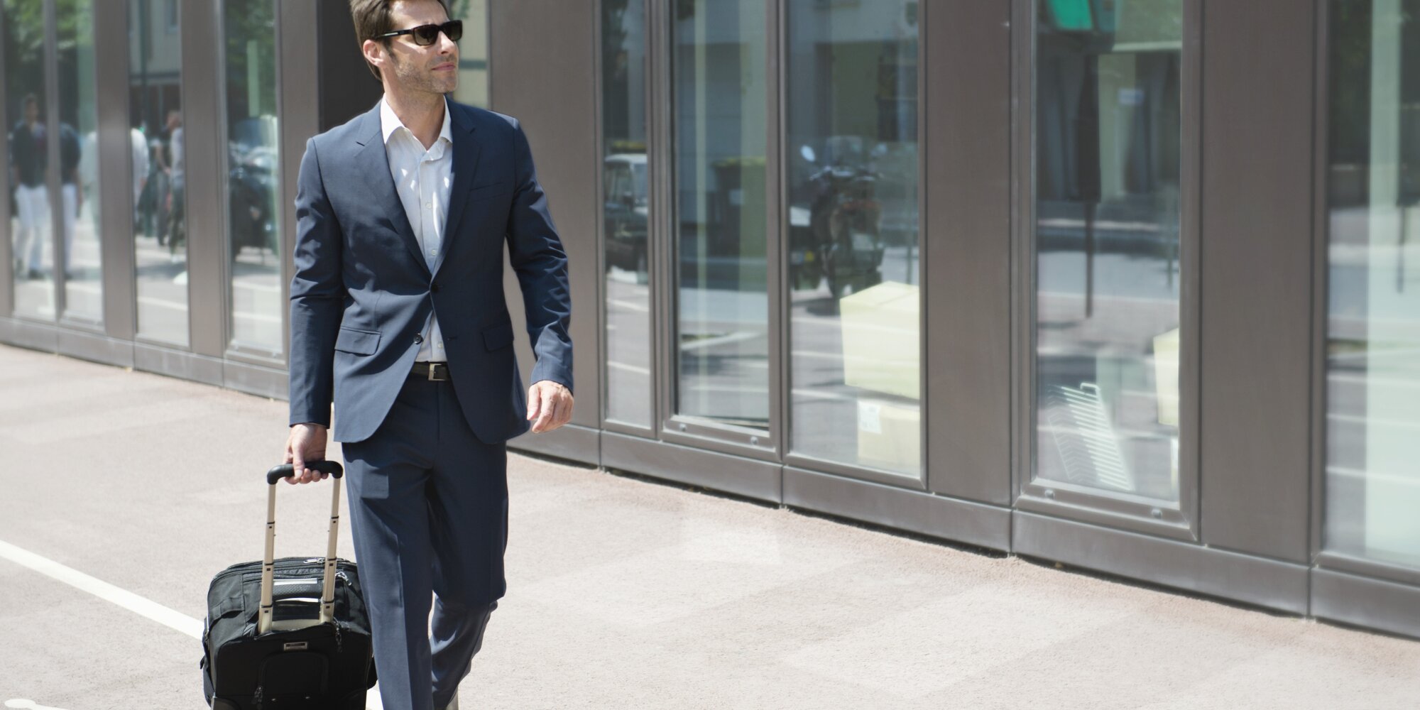 Businessman with sunglasses pulling luggage, walking in city